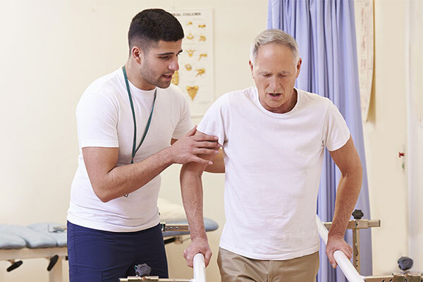 Physical therapist assisting a man using the parallel bars to walk.